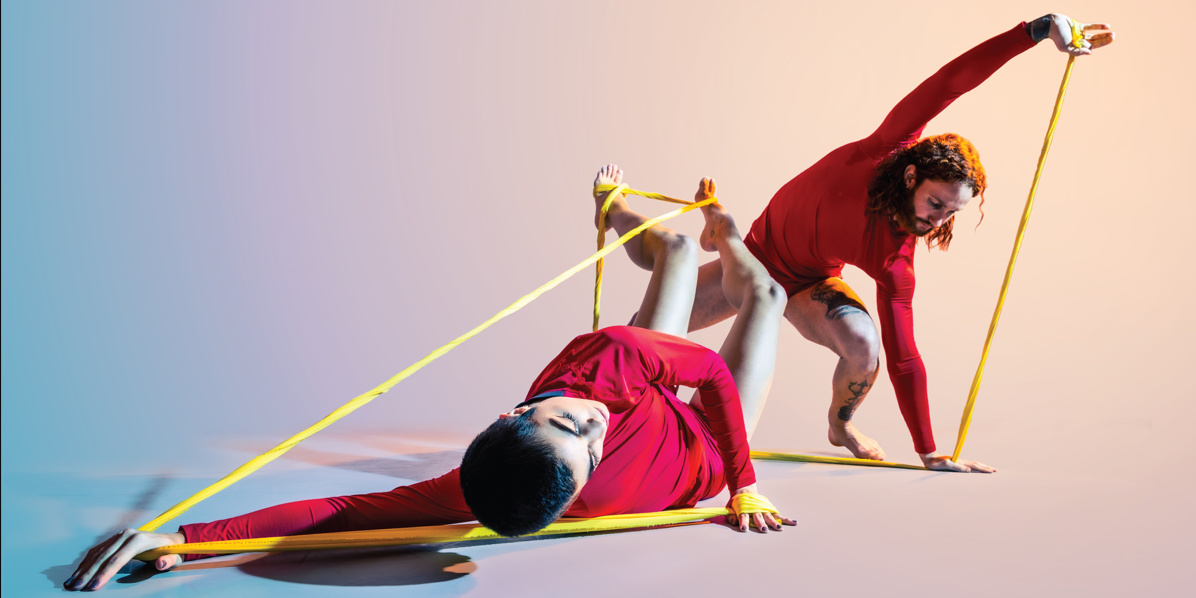 Two dancers in red, exploring movement possibilities when constrained by yellow stretch bands. A conceptual exploration of two new dance works within FIELD THEORY